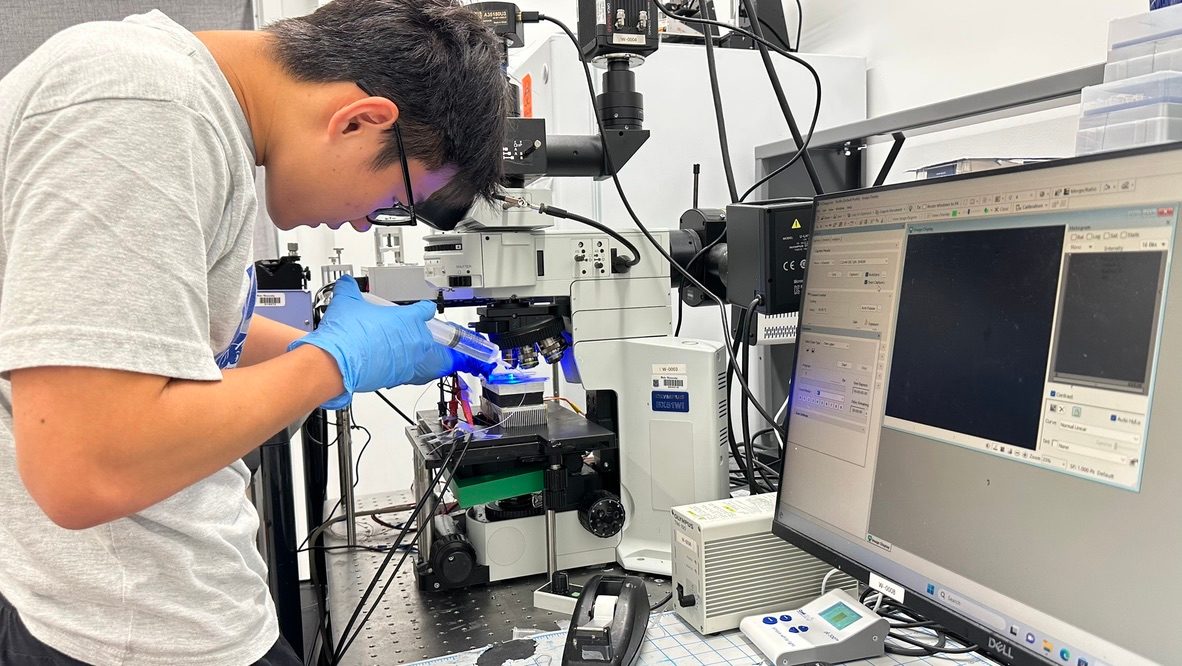 Junhao in nitrile gloves leaning over an optical microscope in the Duke acoustofluidics lab