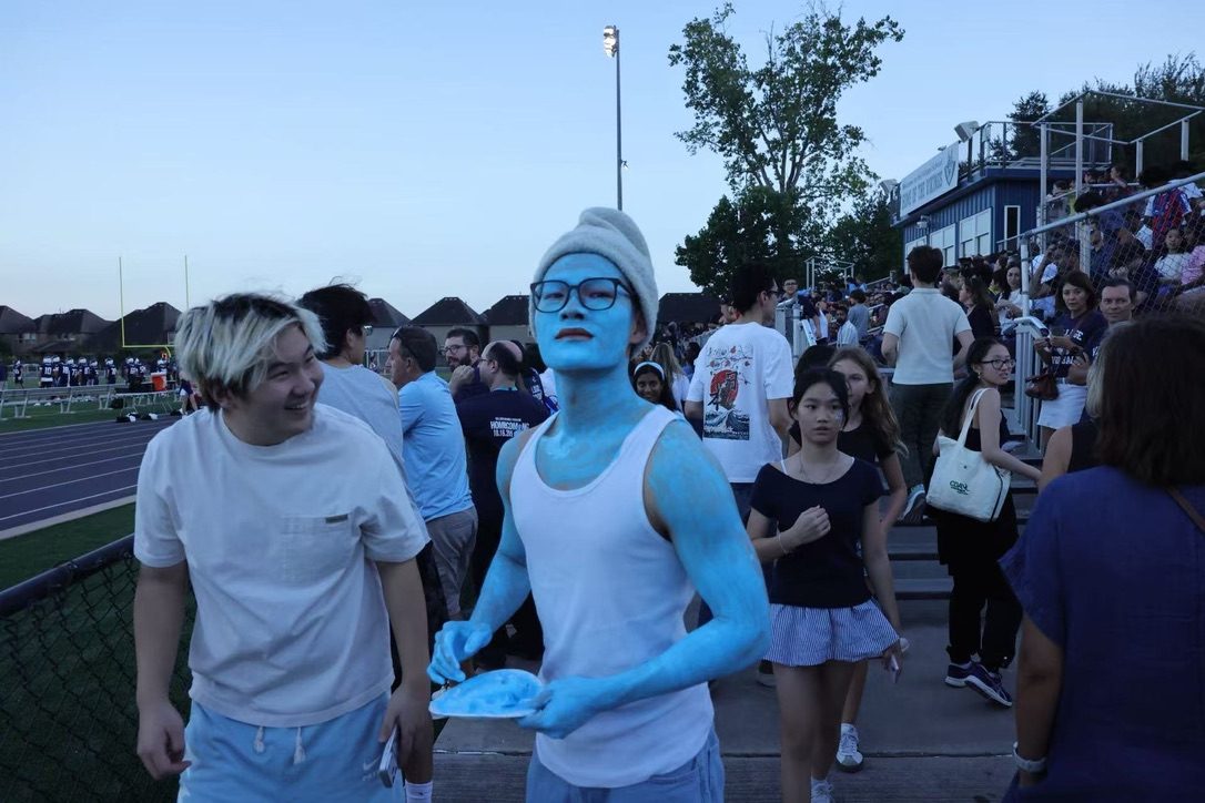 Junhao with blue face and body paint, a Smurf cap and glasses, in a crowd at a school game, with a friend smiling beside him