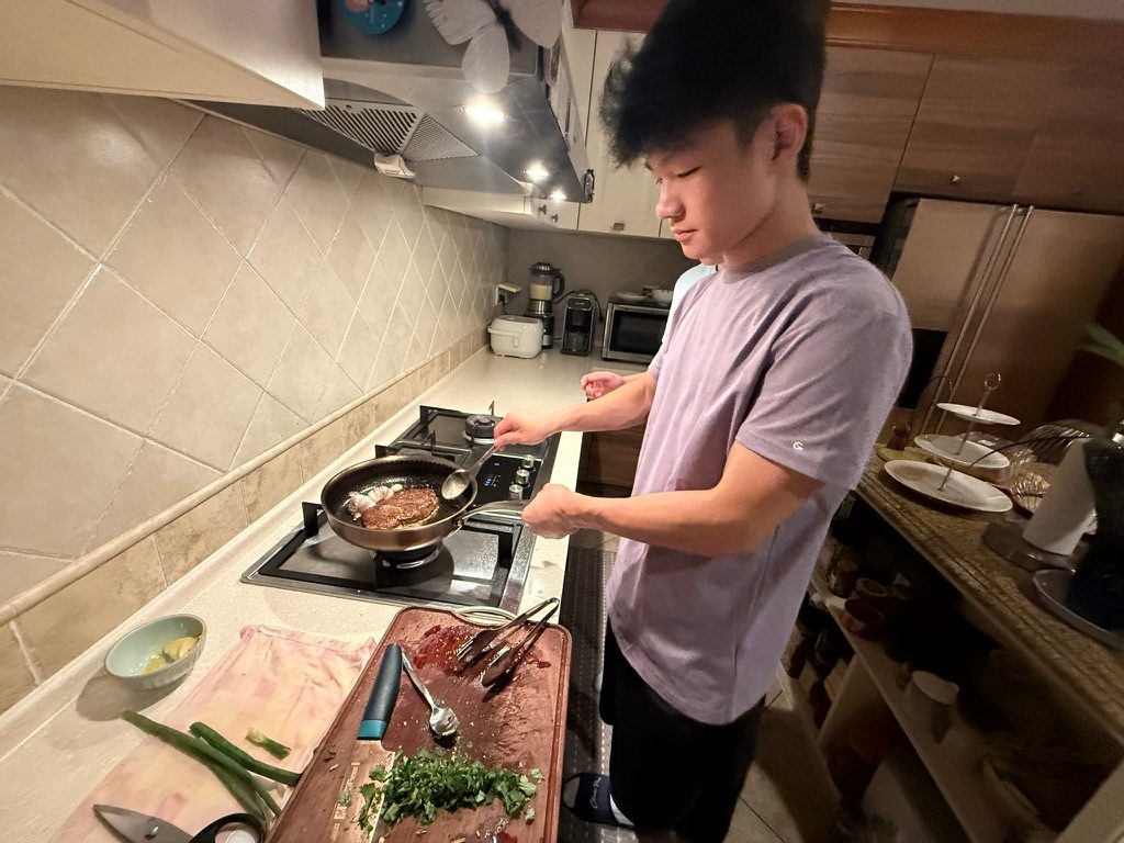 Junhao cooking at a stove, with chopped herbs on a board beside him
