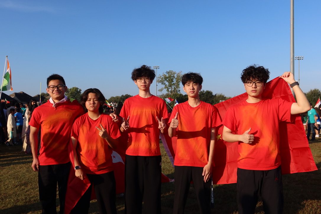 Five friends in matching red shirts, two holding a red cloth like a cape, at an outdoor event