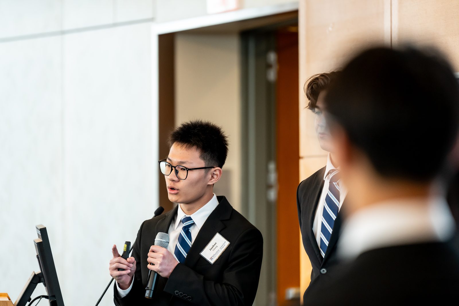 Junhao in a suit speaking into a microphone while presenting at the Wharton Global High School Investment Competition finale